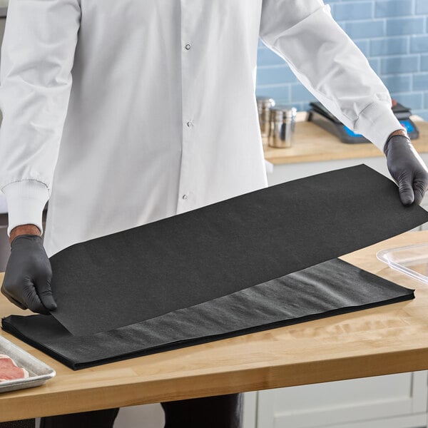 A stack of large black steak paper sheets being handled by a person wearing gloves and a white coat.