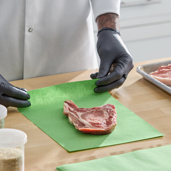 A person wearing black gloves is placing a raw steak on a green 9" x 12" steak paper sheet.