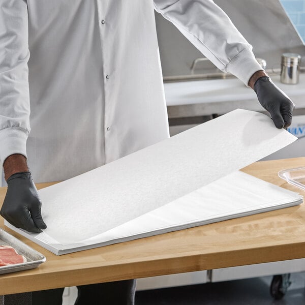 A stack of white 10" x 30" steak paper sheets being handled by a person wearing black gloves in a food preparation area.