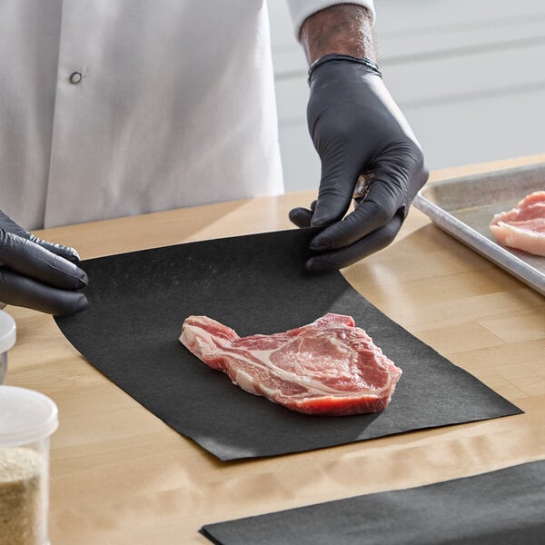 A raw steak placed on a black paper sheet, being handled by a person wearing black gloves.