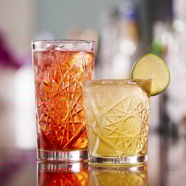 A pair of intricately patterned glass tumblers, one filled with a red beverage and the other with a yellow beverage garnished with a lime slice.
