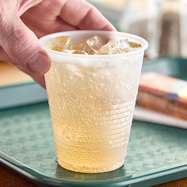 A hand holding a Dart translucent plastic cup filled with ice and liquid on a tray.