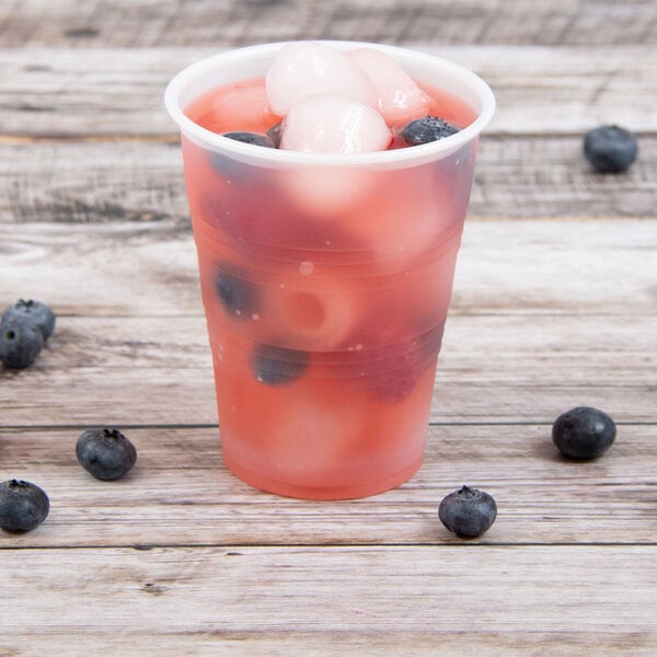 A Dart Conex translucent plastic cold cup filled with blueberry juice and ice with blueberries on a wood surface.