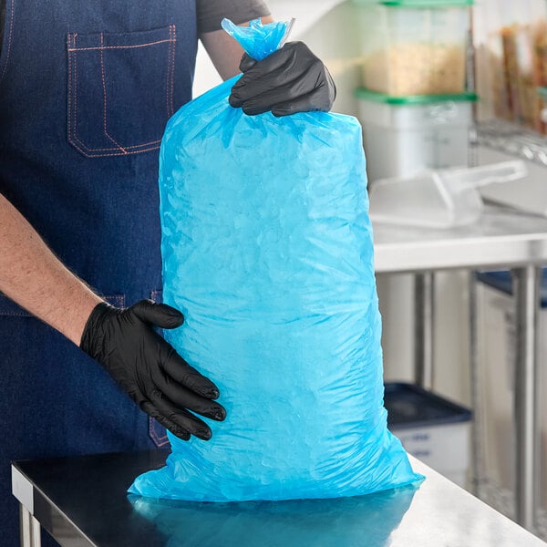 A large blue heavy duty plastic ice bag being held by a person wearing black gloves in a commercial kitchen setting.
