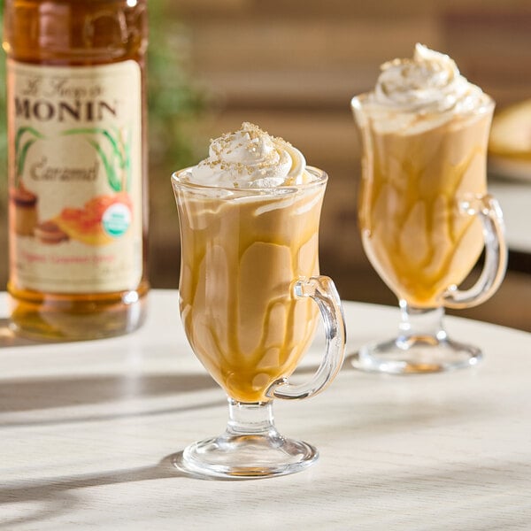 A glass mug filled with a caramel-flavored beverage topped with whipped cream, with a bottle of Monin Organic Caramel Flavoring Syrup in the background.