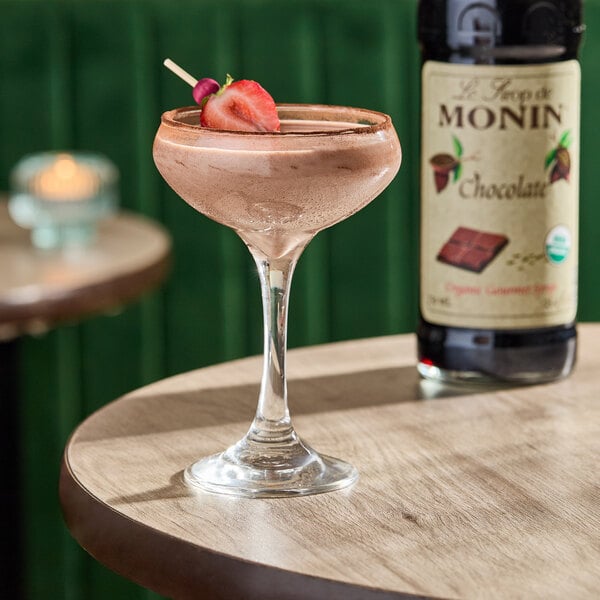 A bottle of Monin Organic Chocolate Flavoring Syrup, 750 mL, is shown next to a chocolate cocktail garnished with a strawberry.
