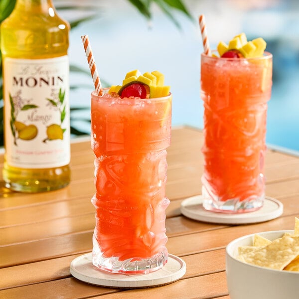 A bottle of Monin Premium Mango Flavoring Syrup next to two tall glasses of a bright orange tropical drink garnished with fruit and a cherry.