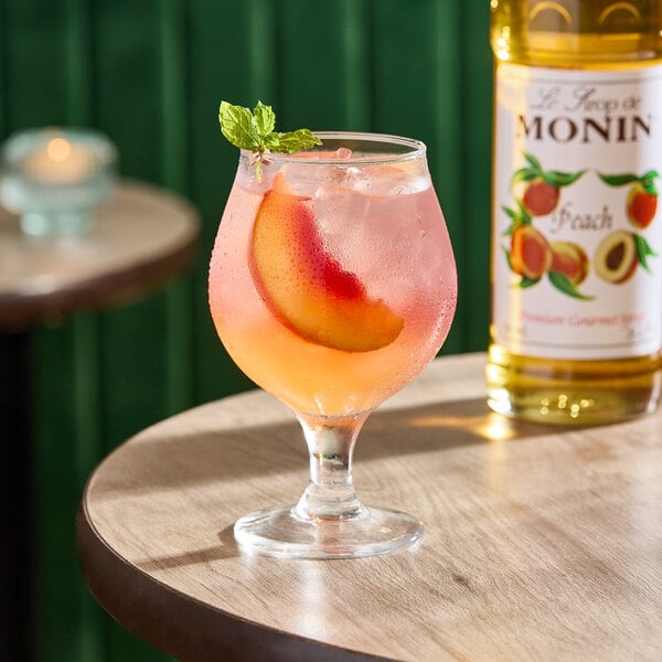 A glass of peach-flavored drink with ice, a peach slice, and a mint garnish, next to a bottle of Monin Premium Peach Syrup.