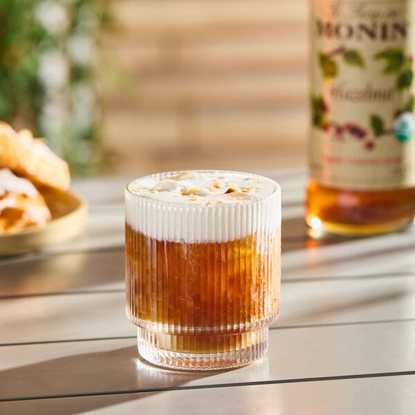 A glass of iced coffee topped with foam, with a bottle of Monin Organic Hazelnut Flavoring Syrup in the background.