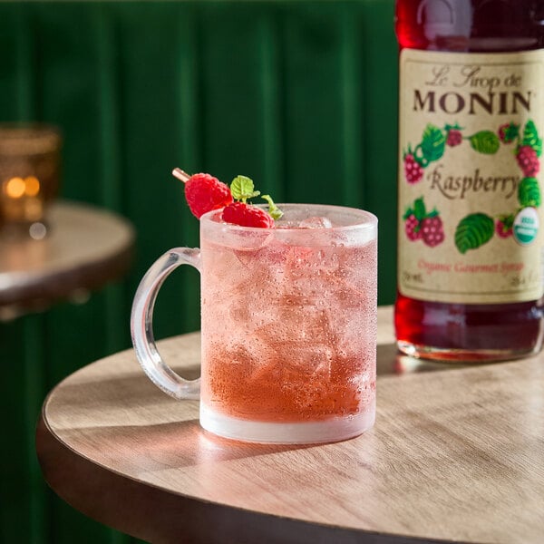 A glass mug filled with a pink raspberry-flavored iced drink, garnished with raspberries and mint, next to a bottle of Monin Organic Raspberry Syrup.