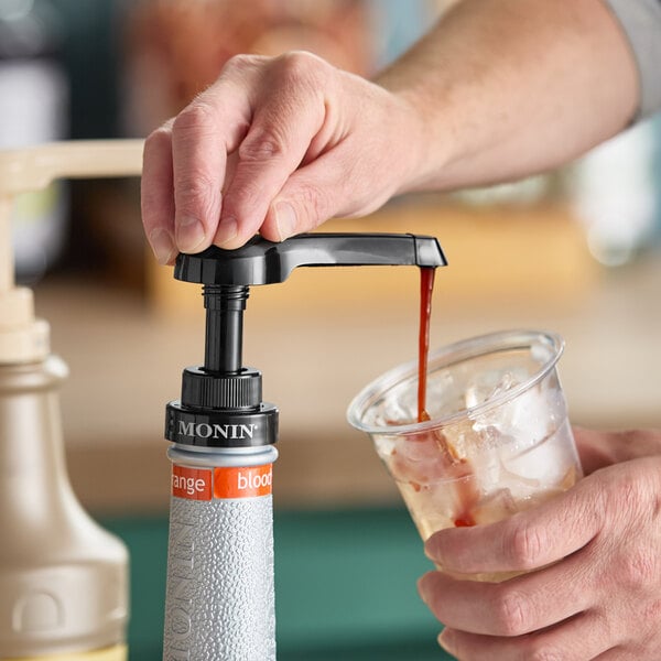 A hand pressing a Monin puree pump to dispense syrup into a plastic cup with ice.