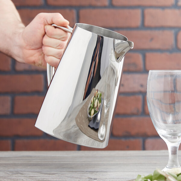 A hand holding a Vollrath stainless steel water pitcher over a glass.