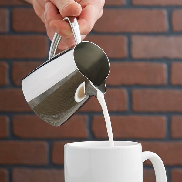 A person pouring milk from a Vollrath stainless steel creamer into a white mug.