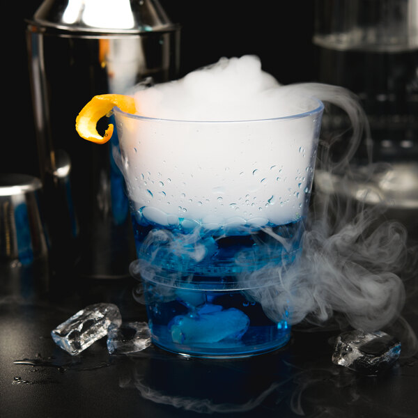 A clear WNA Comet plastic rocks glass filled with a blue drink and ice with smoke and a slice of orange on top.