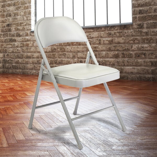 A gray National Public Seating folding chair with gray padding in front of a brick wall.