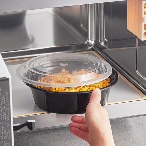 A hand placing a Choice round black plastic container of food in a microwave.