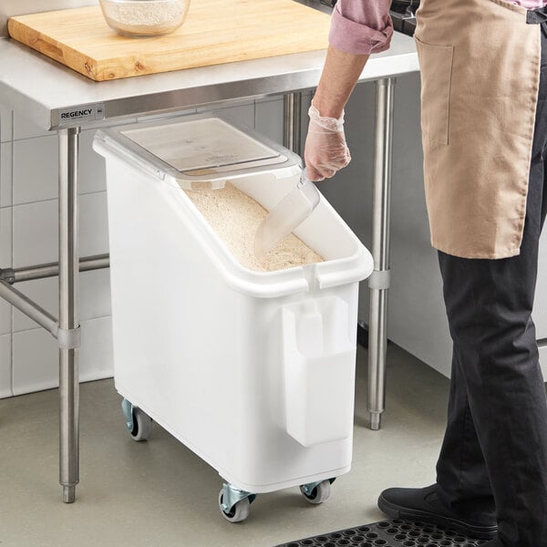 A white slant top mobile ingredient storage bin with a sliding lid and wheels, partially filled with a dry ingredient.
