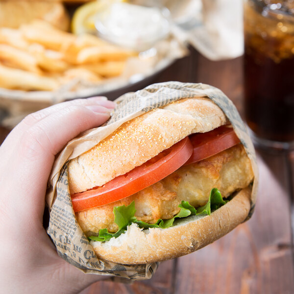 A hand holding a sandwich wrapped in American Metalcraft newspaper print deli paper.