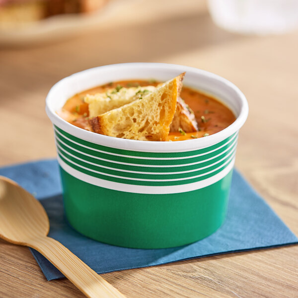 An 8 oz. green paper food cup filled with soup and bread pieces, placed on a blue napkin next to a wooden spoon.