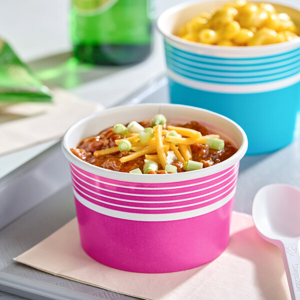 A pink paper food cup filled with chili, topped with shredded cheese and green onions.