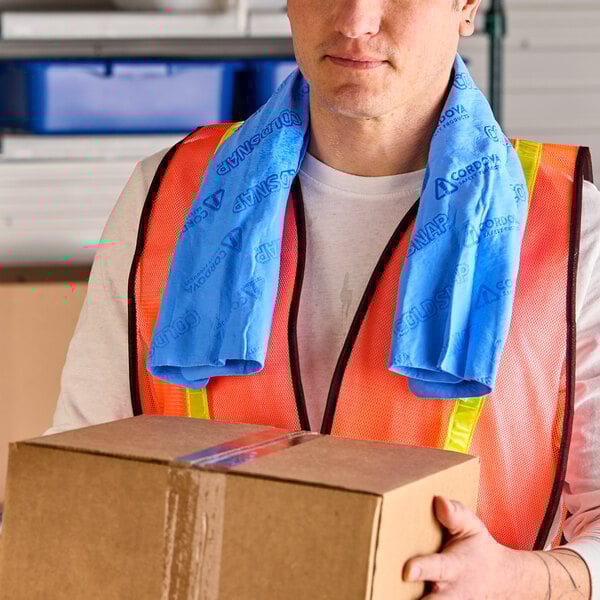 A blue Cordova Coldsnap cooling towel draped around the neck of a person wearing a safety vest and holding a cardboard box.