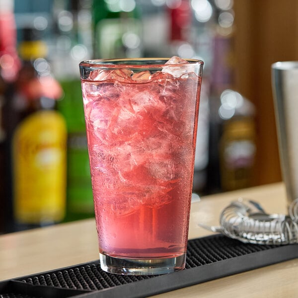 A clear 16 oz. pint glass filled with a red iced beverage, shown on a bar counter.