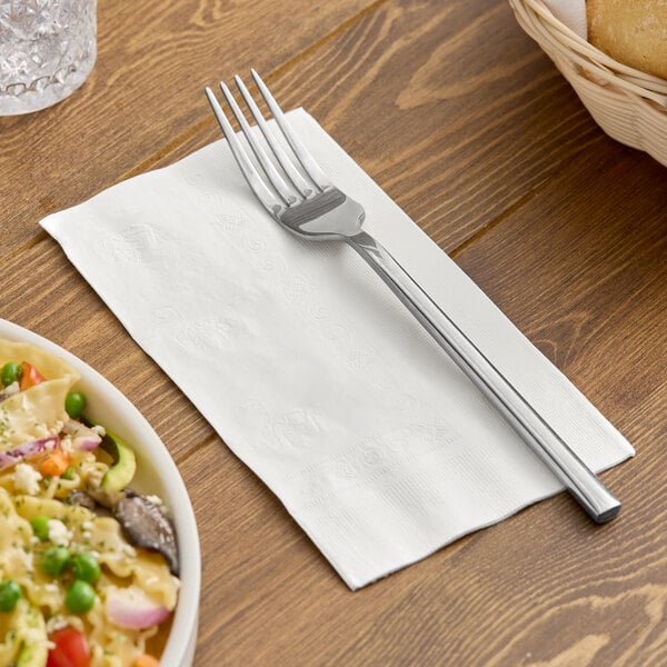 A white 2-ply dinner napkin with a fork placed on top, set on a wooden table.