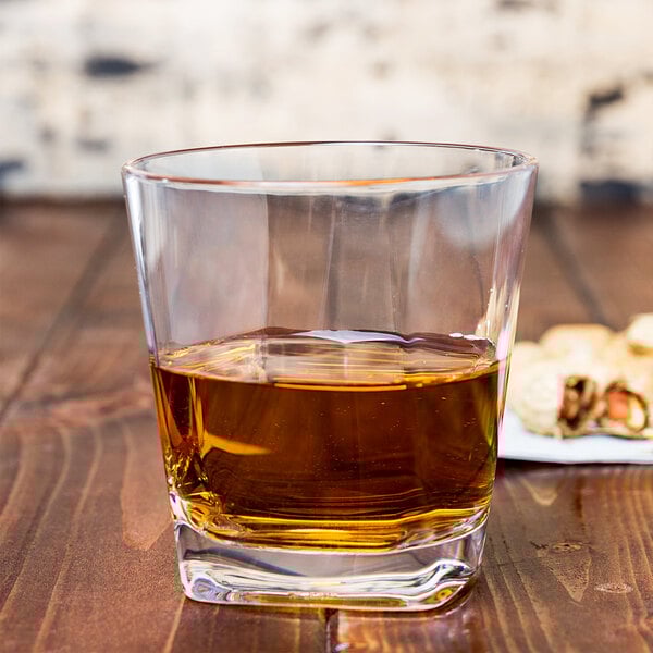 A GET clear plastic rocks glass filled with brown liquid on a table next to a plate of peanuts.