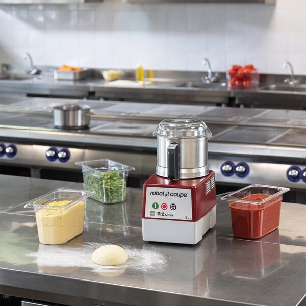 A commercial food processor with a red and white base, clear bowl, and continuous feed chute.