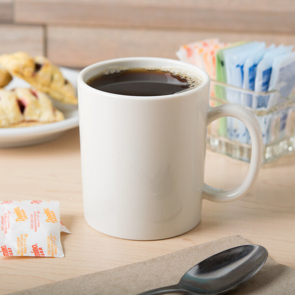 An ivory GET Tritan mug filled with brown liquid on a table with a spoon and a plate of pastries.