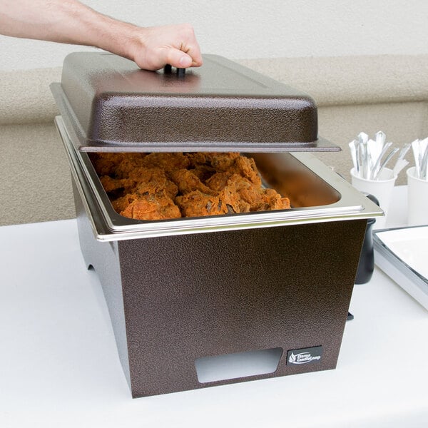 A person holding a Sterno chafer full of food on a table outdoors.