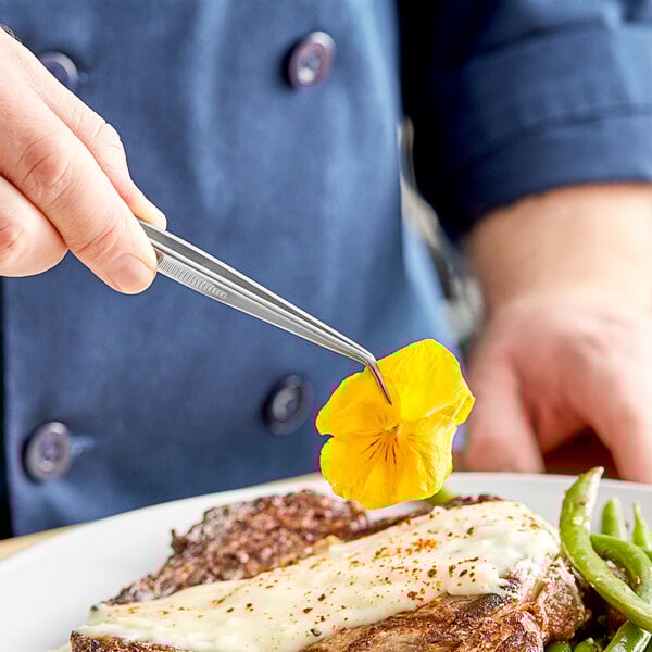 A person holding a pair of Mercer Culinary curved fine point tongs to plate food.