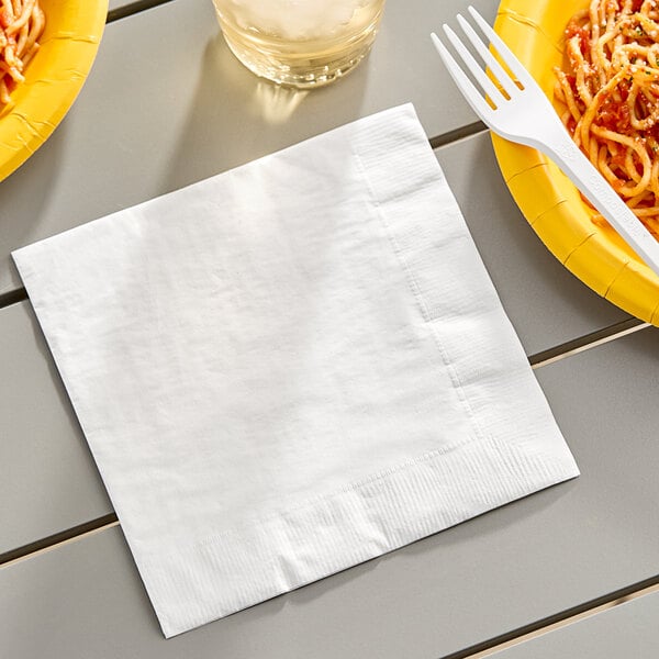 A white Creative Converting 3-ply paper dinner napkin on a table with a plate of spaghetti and a fork.