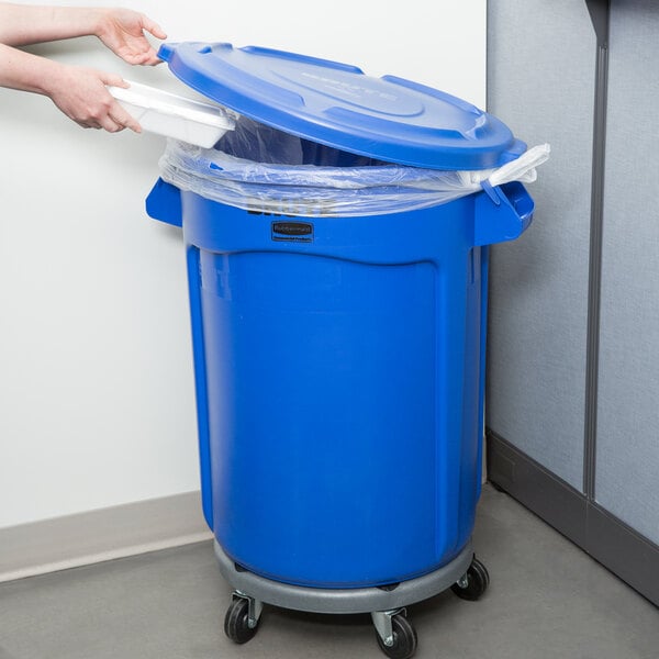 A person holding a blue Rubbermaid BRUTE trash can with a blue lid.