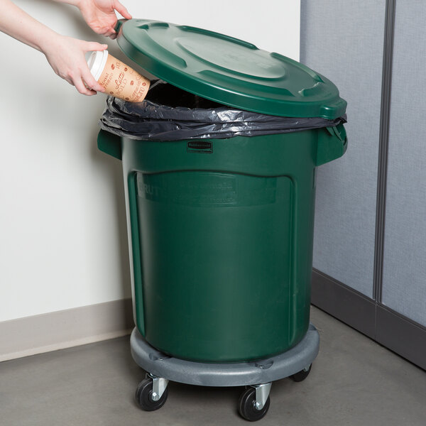 A hand putting a coffee cup into a green Rubbermaid BRUTE trash can.