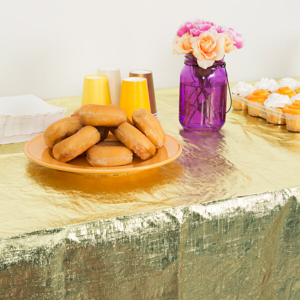 A gold metallic plastic table cover on a table with doughnuts and cupcakes.