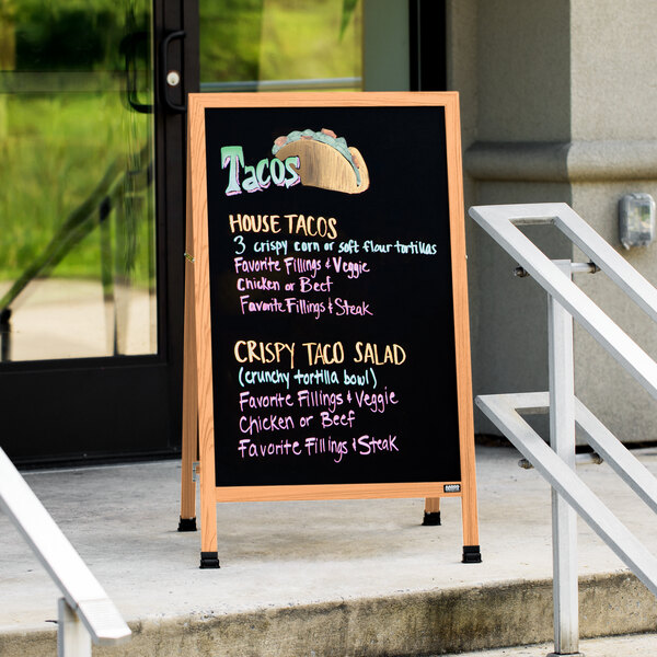 An Aarco solid oak A-frame sidewalk board with a black porcelain marker board on a sidewalk.