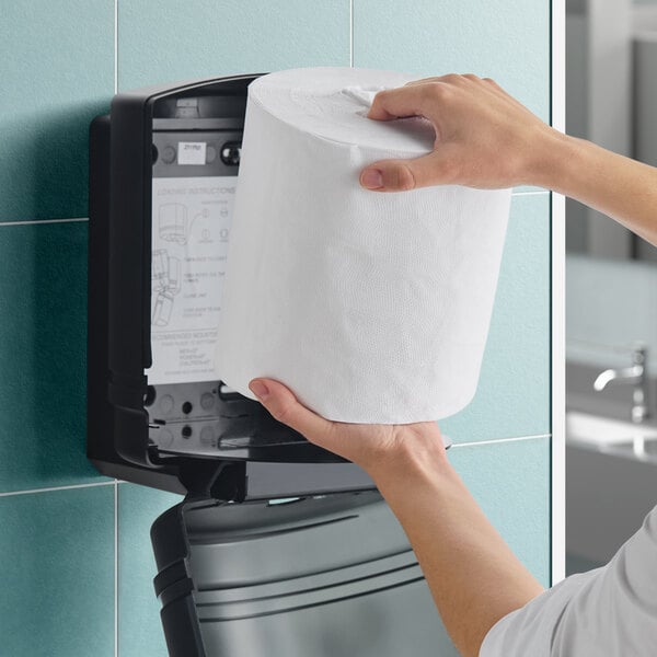 A person loading a large white center pull paper towel roll into a black wall-mounted dispenser.