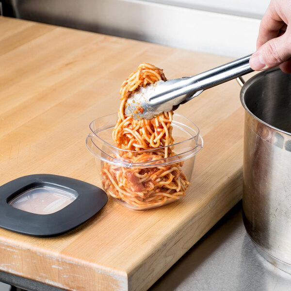 A person using tongs to serve spaghetti from a Rubbermaid clear square food storage container.