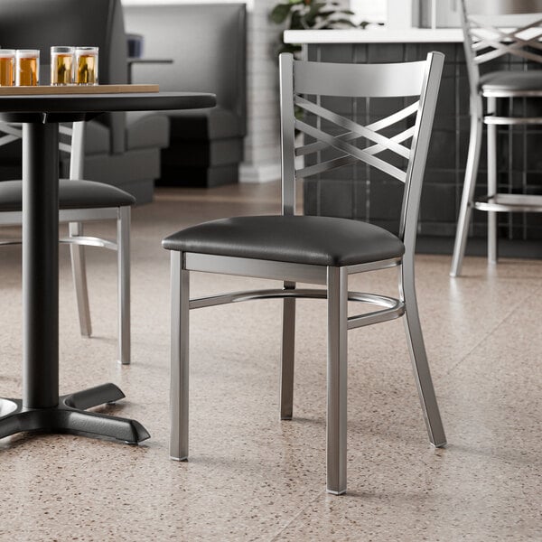 A metal cross back chair with a clear coat finish and a black vinyl padded seat.