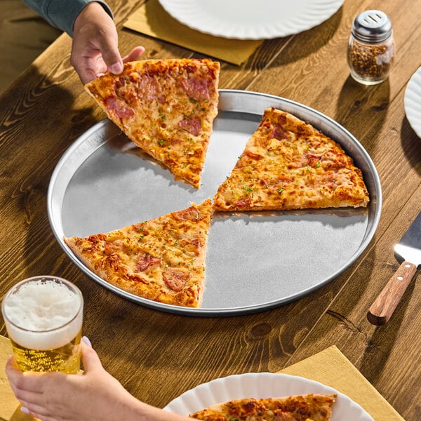 A round, silver aluminum pizza pan with a low rim, holding three large slices of pizza on a wooden table.