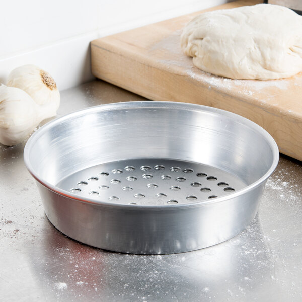 An American Metalcraft silver pizza pan with holes in it holding dough.