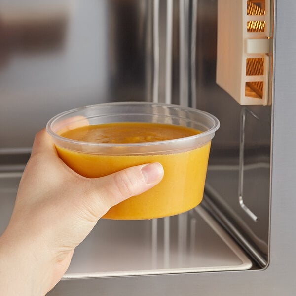 A hand holding a Choice translucent round deli container of food in front of a microwave.