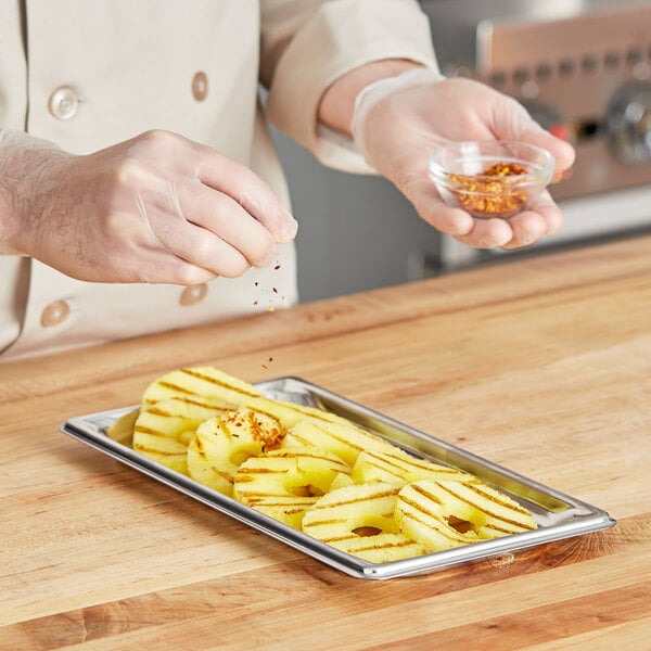 A person pouring seasoning onto a tray of pineapple slices using a Vollrath stainless steel steam table pan.