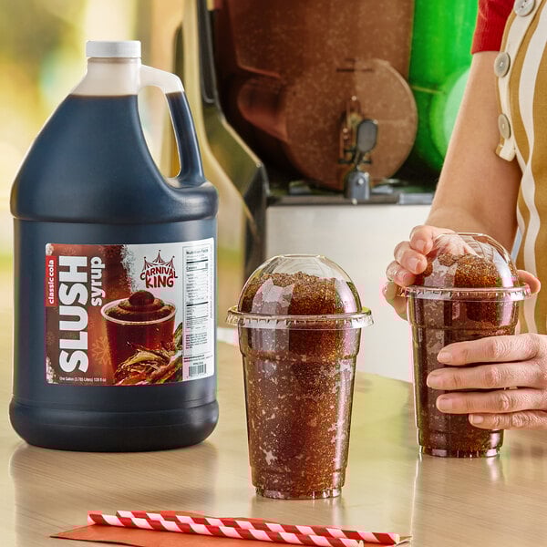 A gallon jug of Carnival King cola slush syrup concentrate next to two cups of prepared cola slushies with dome lids.