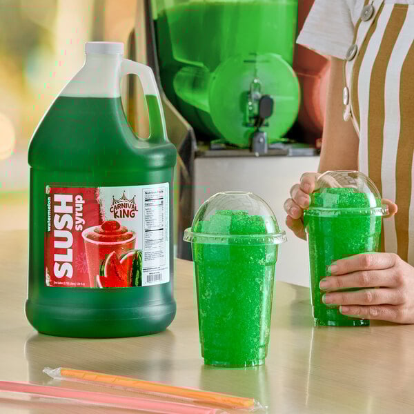 A gallon jug of Carnival King watermelon slush syrup concentrate next to two cups of green slushy drinks.