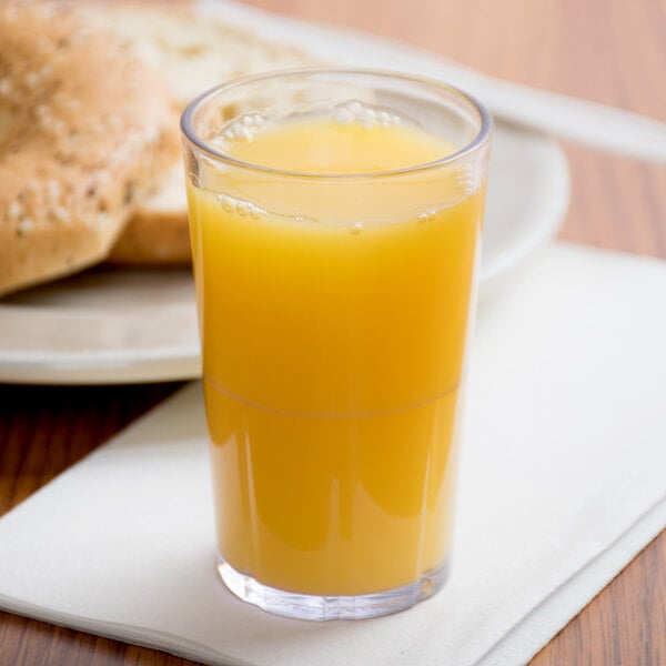 A clear plastic Cambro tumbler filled with orange juice on a table next to a bagel