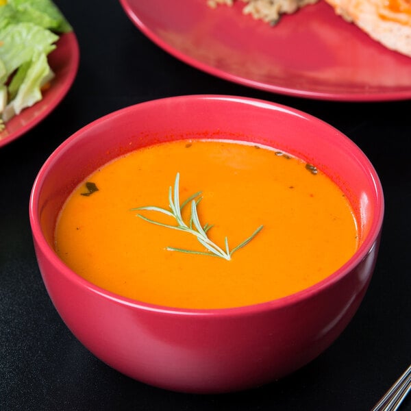 A red 10 Strawberry Street Wazee Matte stoneware bowl filled with soup garnished with a sprig of rosemary.