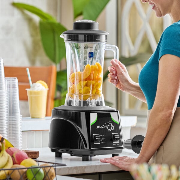 A commercial blender with a clear 64 oz. container filled with fruit, shown on a countertop with a person operating it.