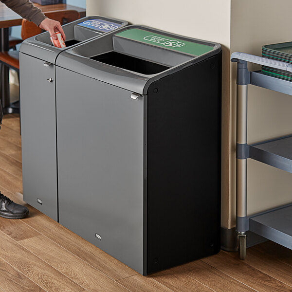 A person standing next to a grey Rubbermaid rectangular recycling container with a green organic waste label.